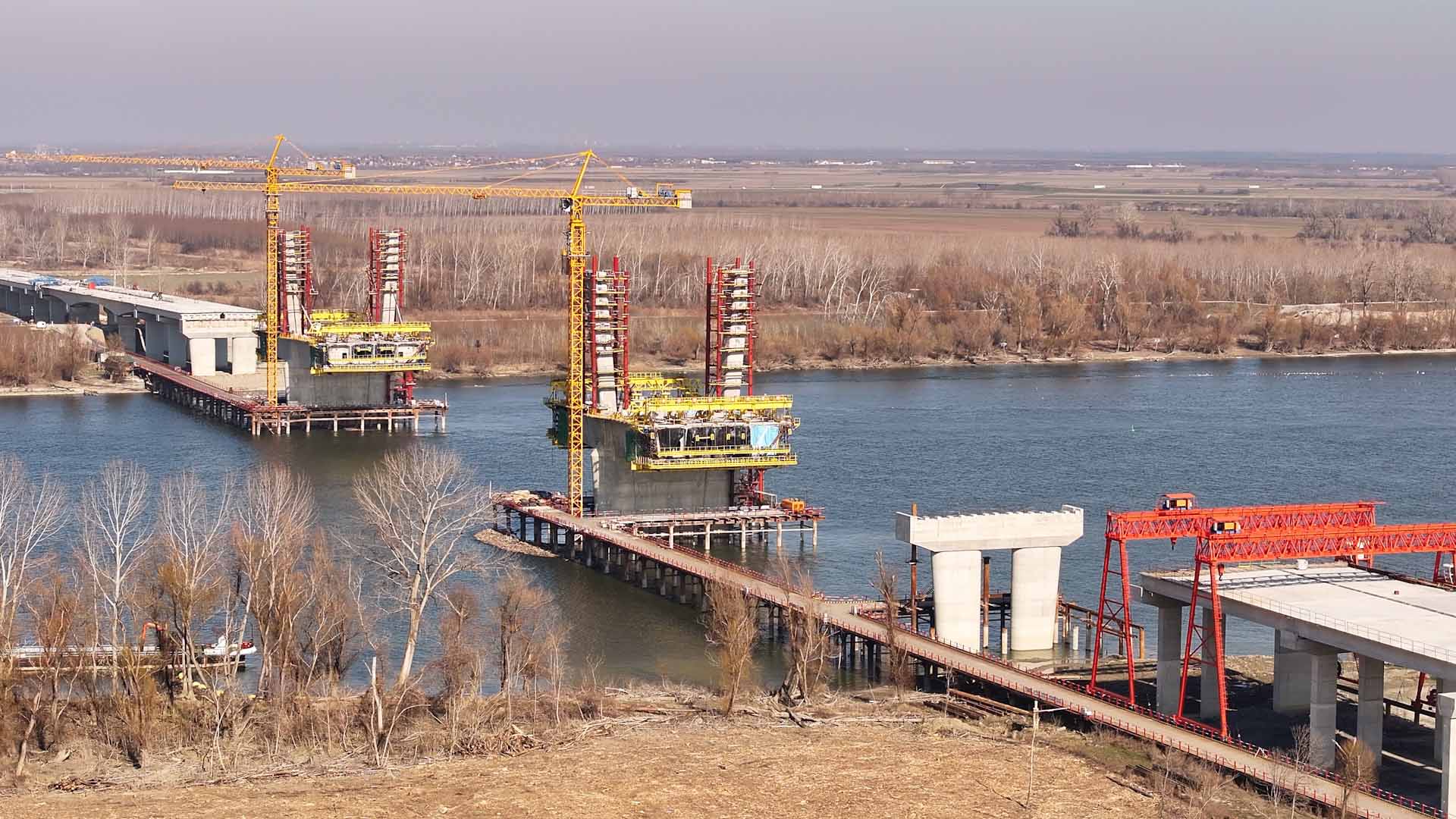 "Izgradnja petog novosadskog mosta u sklopu Fruškogorskog koridora, betonski stubovi i Dunav u pozadini."